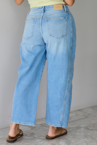A woman wearing light-wash blue cropped barrel cut jeans with an asymmetrical button closure, standing indoors against white wall with white marble flooring. The photo shows the back of the jeans, which have functional back pockets. 