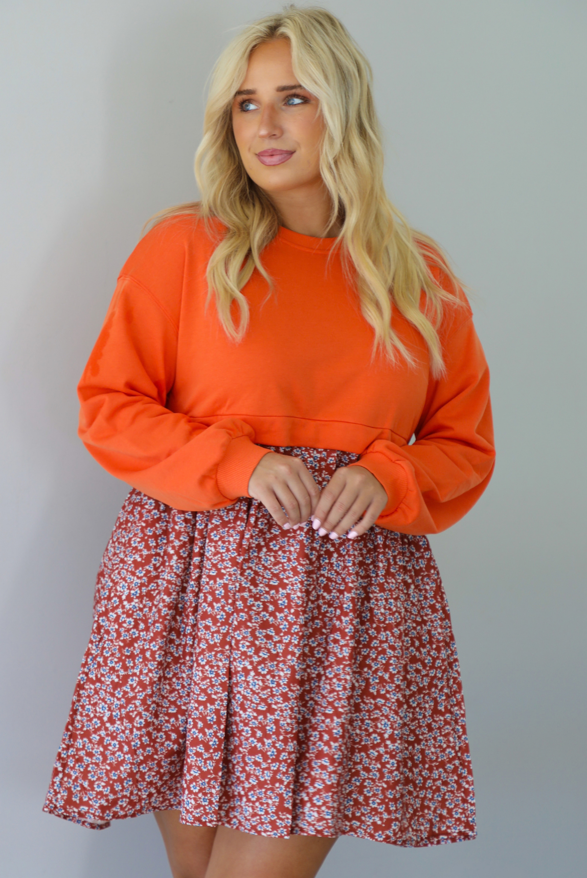 A woman wearing a block style oversized, flowy dress featuring a solid orange bodice and sleeves with a multi-colored floral skirt, standing indoors against a white wall.
