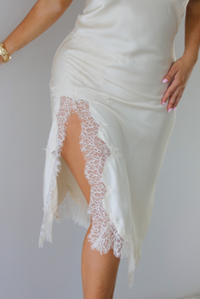 Woman wearing an off white satin midi length dress that has spaghetti straps, lace detailing at the v-neckline, and lace detailing at the hem and split hem detail on the right side. The photo is taken indoors against a white wall and shows a closeup of the split hem design on the right leg with its lace detailing. 