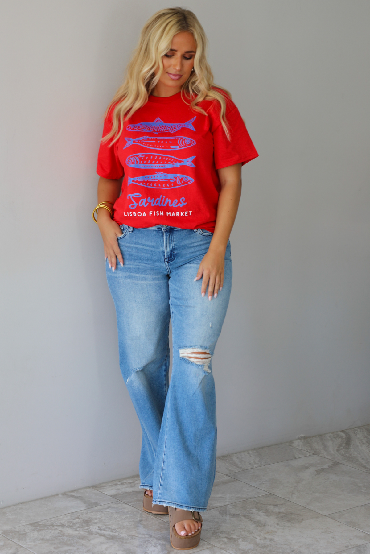 A woman wearing a red graphic t-shirt with blue sardine illustrations and the text 'Sardines LISBON FISH MARKET', paired with blue jeans, standing indoors against a white wall with white marble flooring.