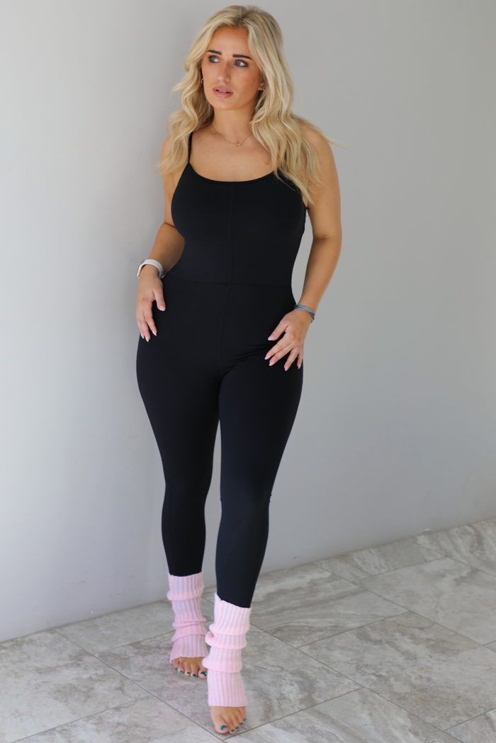 Woman wearing a black fitted jumpsuit with spaghetti straps. The jumpsuit is styled with pink leg warmers. The photo is taken indoors against a white wall with white marble flooring. 