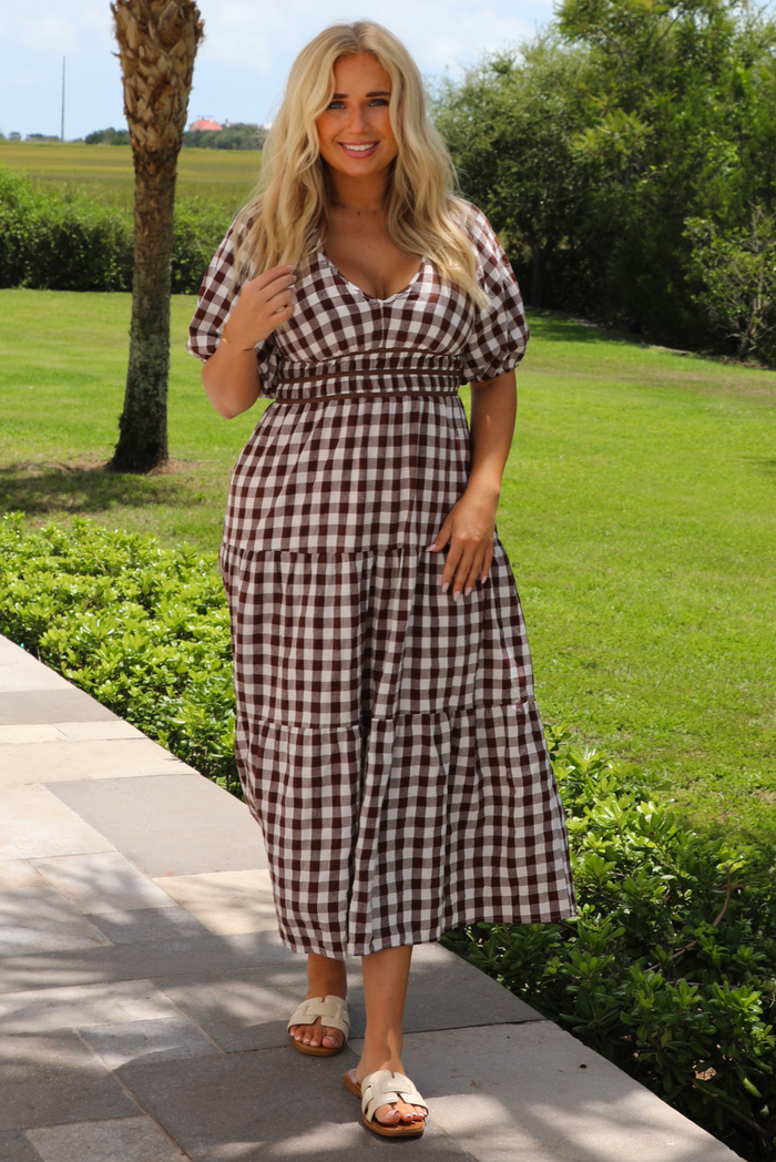 A woman wearing a brown and white gingham tiered midi dress with puff sleeves, standing outdoors on a stone pathway with green grass and trees in the background.