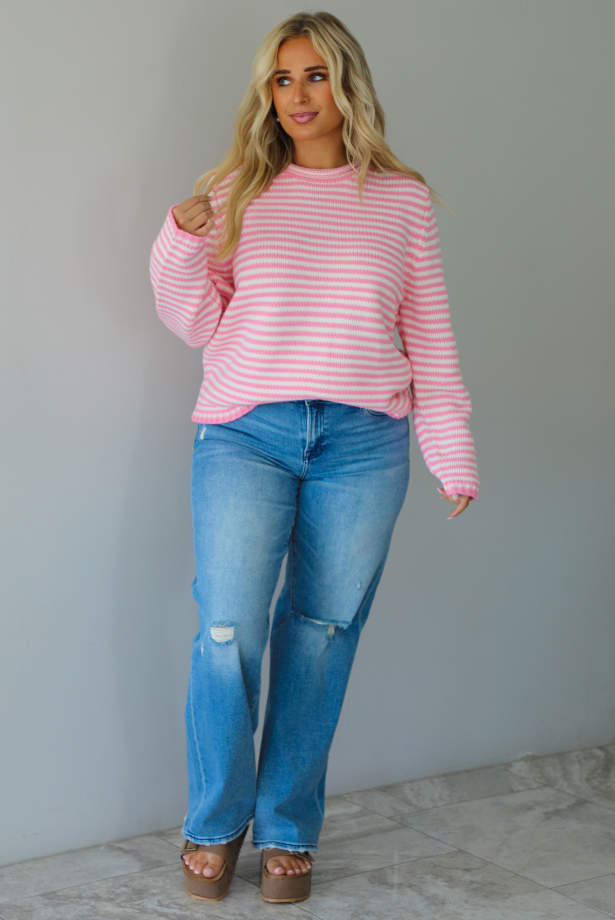 A woman wearing a pink and white striped sweater paired with light wash denim jeans, standing indoors against a white wall with white marble flooring. 