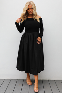 Woman wearing a black off the shoulder, long sleeved midi length dress with a pair of clear strapped nude heels. The photo is taken outdoors against a white wall.