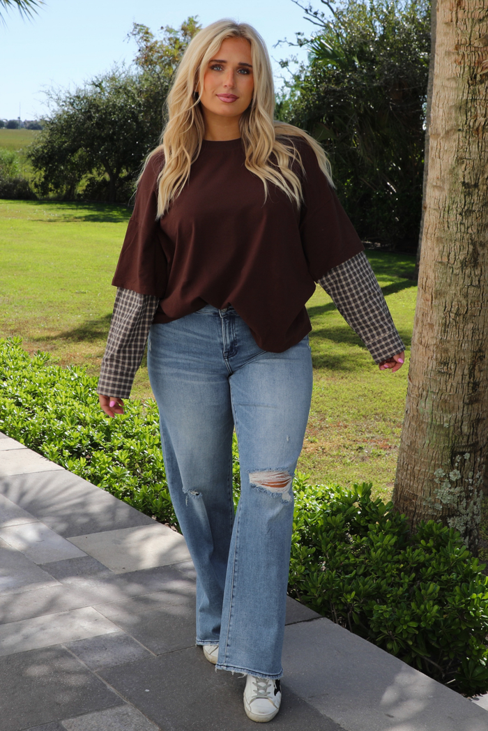 A woman wearing a brown t-shirt with a layered look of plaid sleeve detail, paired with light wash denim jeans, standing outdoors on a stone path with palm trees and other greenery in the background. 
