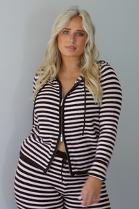 A woman wearing a light pink and brown striped two piece set featuring a double zipped hoodie top and mid-rise, wide leg pants, standing indoors against a white wall. The photo shows a closeup of the top and how the top can be zipped from the top or bottom. 