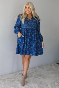 A woman wearing a dark denim mini dress with a white floral print pattern throughout, collared neckline, a quilted detail at the bodice, long sleeves, and a flowy skirt, standing indoors against a white wall with white marble flooring. 