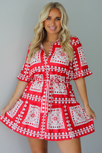 A woman modeling a red and white patterned short-sleeve mini dress with button closures along the front and a tie waist, standing indoors against a white wall.