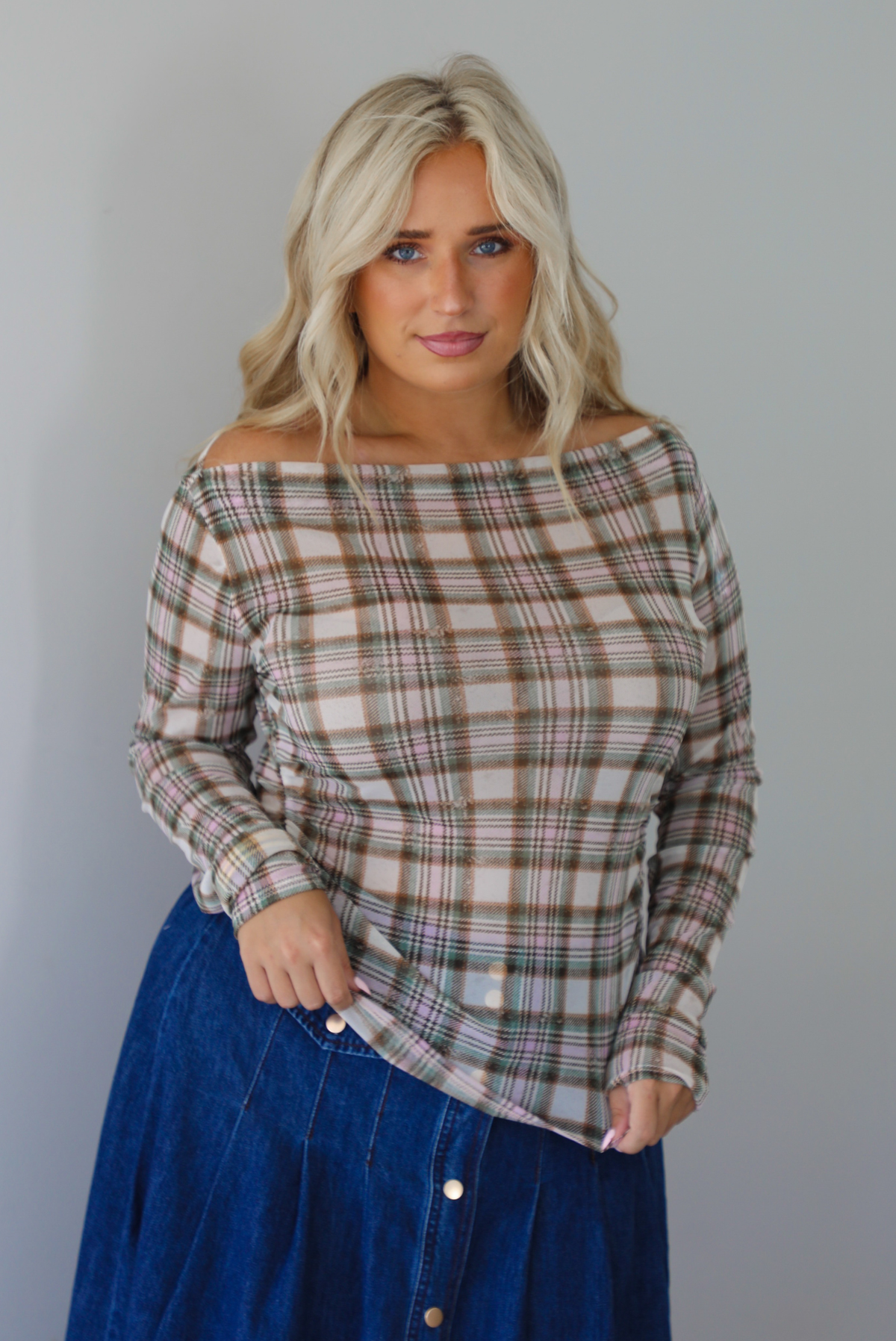 A woman modeling an off-the-shoulder long-sleeve plaid top in beige, brown, and green tones, paired with a blue denim skirt, indoors against a white wall. 