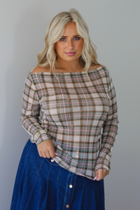 A woman modeling an off-the-shoulder long-sleeve plaid top in beige, brown, and green tones, paired with a blue denim skirt, indoors against a white wall. 