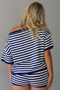 A woman shown from behind wearing a navy and white horizontally striped off-the-shoulder top and matching striped shorts, standing indoors against a white wall.