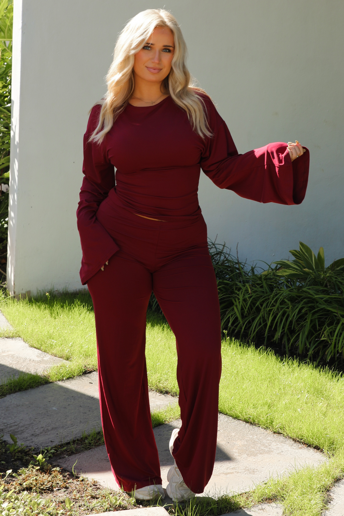 Woman model wearing a burgundy wine colored two piece set outdoors. The set features a slightly cropped, long sleeved top that has flared sleeves and a boat neckline. The pants are a mid-rise, fitted on the thighs, and have a straight cut design at the bottom. It's super soft, lightweight, and stretchy. 