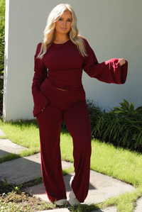 Woman model wearing a burgundy wine colored two piece set outdoors. The set features a slightly cropped, long sleeved top that has flared sleeves and a boat neckline. The pants are a mid-rise, fitted on the thighs, and have a straight cut design at the bottom. It's super soft, lightweight, and stretchy. 