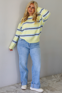 Woman wearing a pastel yellow, blue, and dark brown striped sweater, paired with light wash denim jeans that have a wide leg cut with a cuffed hem, standing indoors against a white wall with white marble flooring.