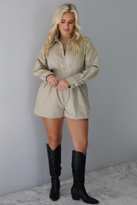 A woman wearing a sand colored long sleeved romper with a collared neckline, functional zipper closure in the front, pleated details throughout, functional pockets on either side, paired with black colored knee-high boots, standing indoors against a white wall with white marble flooring. 