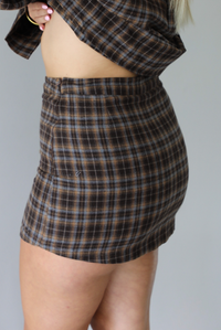 A woman wearing aa dark brown plaid set featuring an oversized button-down and fitted mini length skirt, focusing on the mini skirt showing the zipper closure on the side, standing indoors against a white wall.