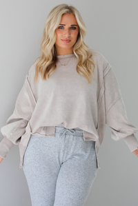 Woman wearing sand colored oversized sweatshirt with exposed seam details and a high-low design with heather grey casual pants. The photo is taken indoors against a white wall.