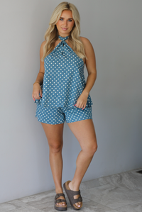 Woman wearing denim blue with white polka dot halter tie top and high waisted shorts indoors against a white wall with white marble flooring. 