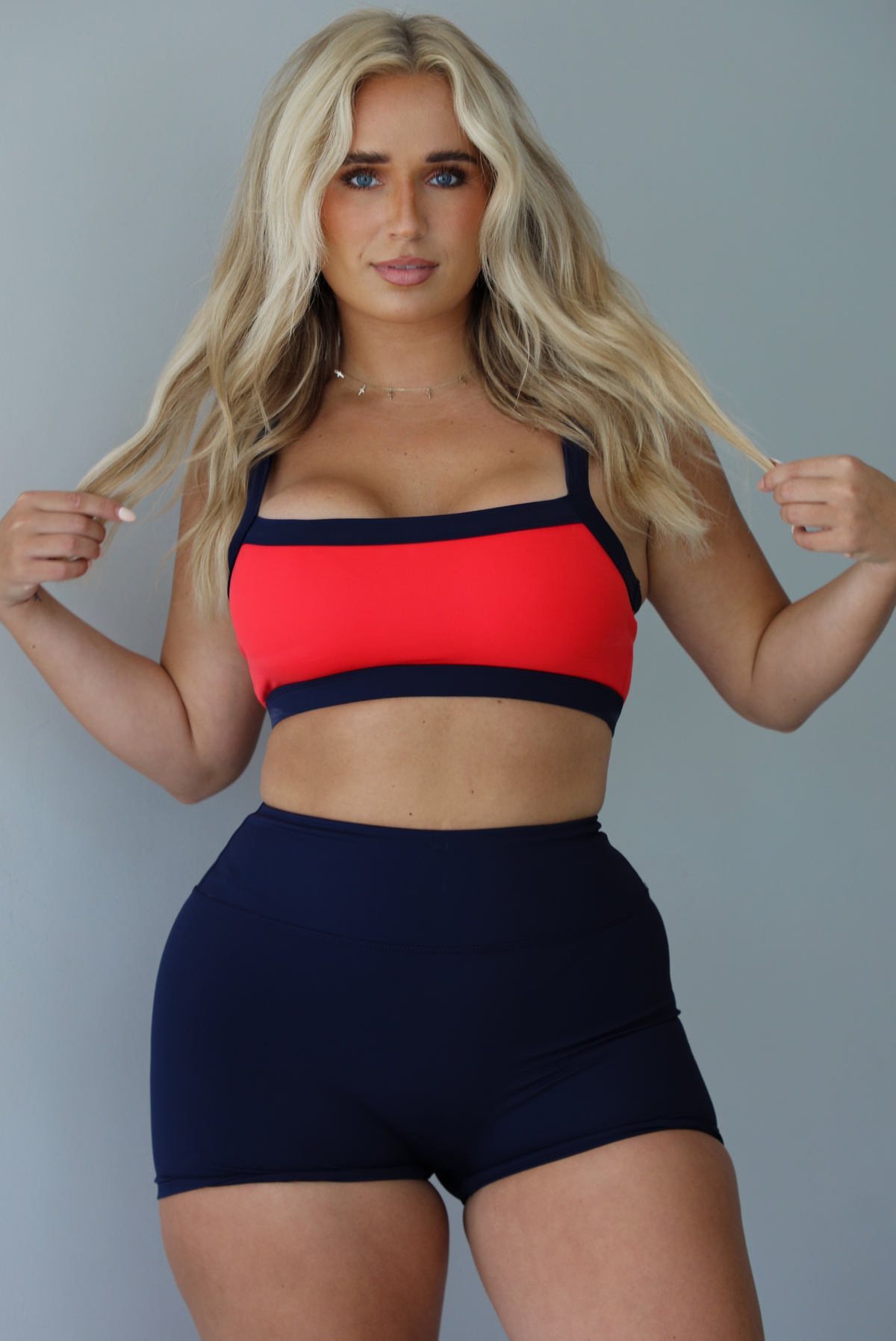 Woman wearing. navy and red sports bra with navy high waisted shorts indoors against a white wall.