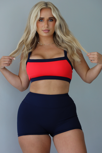 Woman wearing. navy and red sports bra with navy high waisted shorts indoors against a white wall.