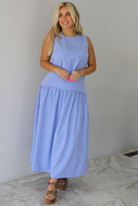 Woman wearing a blue and white gingham two piece set featuring a sleeveless top and high waisted, maxi length skirt indoors against a white wall with white marble flooring. 
