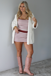 A woman wearing a cream colored oversized cardigan worn over a striped two piece set, paired with knee high pink boots, standing indoors against a white wall with white marble flooring. 