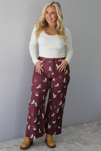 A woman wearing a cream colored knit long sleeved top paired with high waisted maroon jeans with a repeating mallard design, standing indoors against a white wall with white marble flooring. 