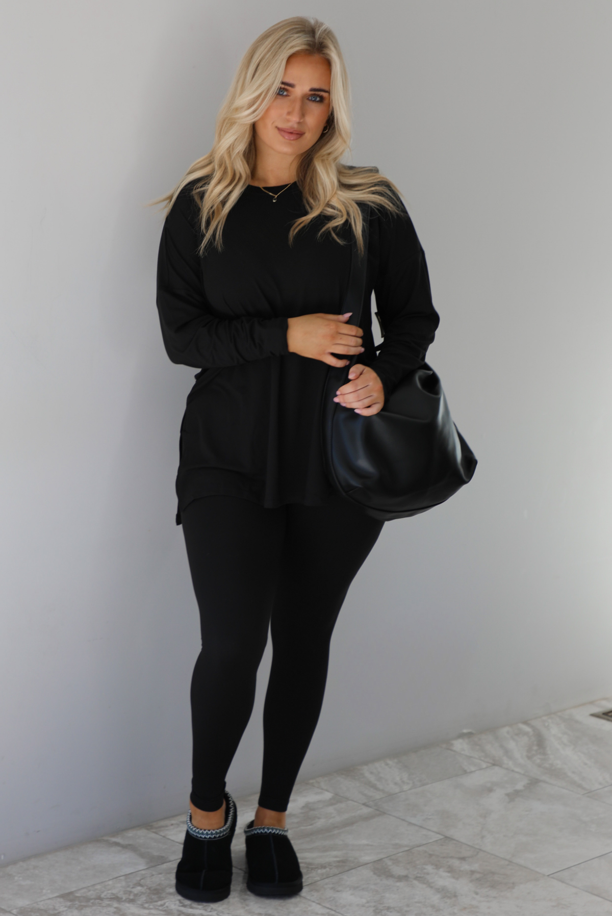 Woman wearing a black two piece set that features an oversized long sleeved top and fitted leggings. The set is paired with a black vegan leather shoulder bag and black slide on slippers. The photo is taken indoors against a white wall with white marble flooring.