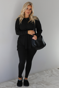Woman wearing a black two piece set that features an oversized long sleeved top and fitted leggings. The set is paired with a black vegan leather shoulder bag and black slide on slippers. The photo is taken indoors against a white wall with white marble flooring.