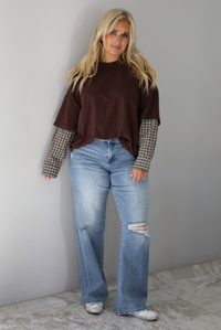 A woman wearing a brown t-shirt with a layered look of plaid sleeve detail, paired with light wash denim jeans, standing indoors against a white wall with white marble flooring. 