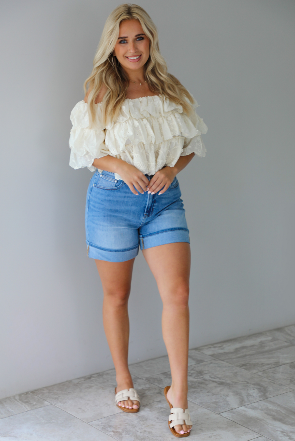 Woman wearing a cream off the shoulder, ruffled eyelet cropped top with medium wash denim shorts indoors against a white wall with white marble flooring. 