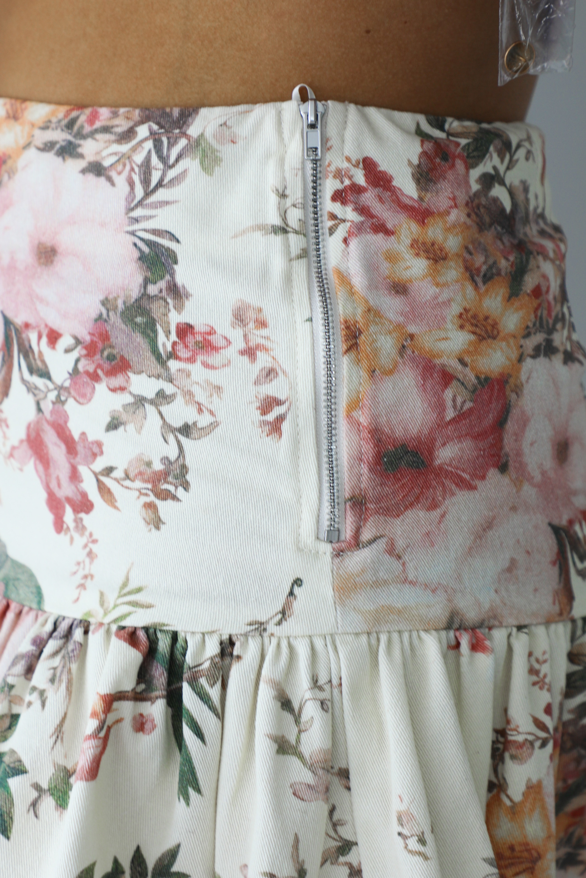 Woman wearing a cream and multi-colored floral skirt that has a high waistline and a shorter length. The photo shows a closeup of the pattern and colored of the skirt, which displays a white variety of flora patterns with branches and birds. It also shows the zipper closure which is located on the left side. 