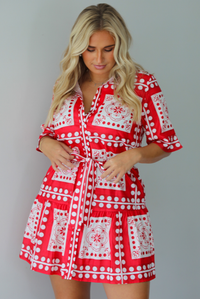 A woman modeling a red and white patterned short-sleeve mini dress with button closures along the front and a tie waist, standing indoors against a white wall.