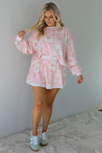 Woman wearing a two piece set featuring a pink floral set that has an oversized long sleeved top and high waisted skirt. The photo is taken indoors against a white wall with white marble flooring. 