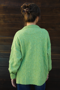 Back of a woman model wearing a green cable knit zip-up cardigan with dark denim jeans outdoors against a dark brown wooden door. The model is wearing a Small. 