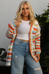Woman model wearing an orange, lavender, yellow, muted green, blue, and white striped oversized cardigan with functional button closures in the front and an oversized fit with a light wash flared hem denim. The photo is taken outdoors against a white wall and greenery in the back. The photo showcases a close up of the cardigan unbuttoned and shows the full length, which falls to the upper thigh on our 5'7" model.