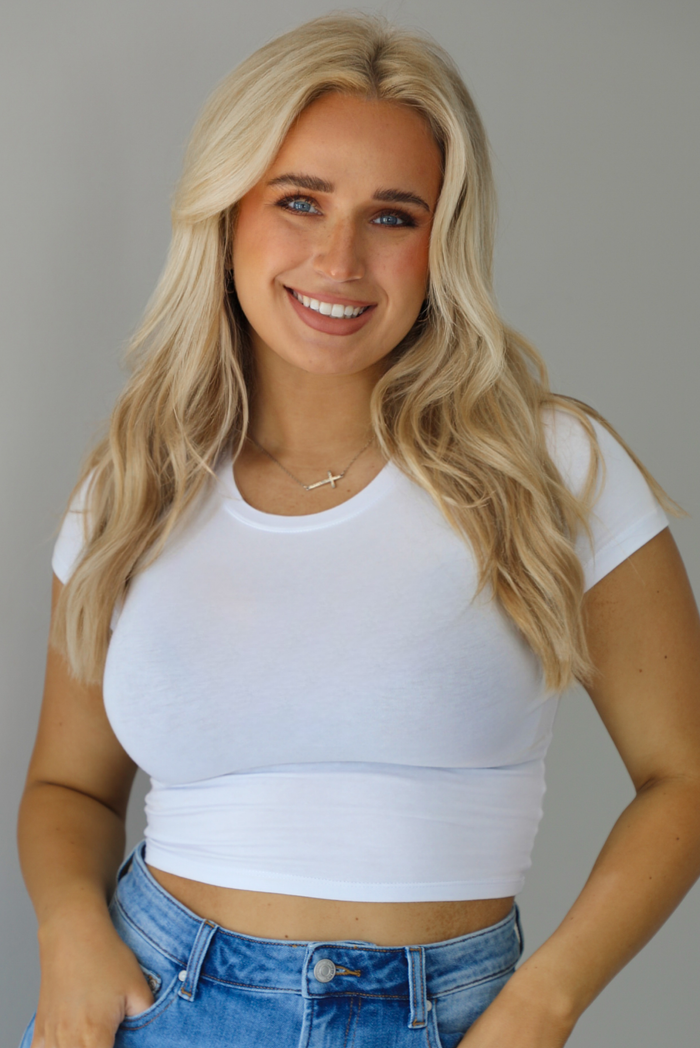 Woman wearing a white, short sleeved crop top. The photo is taken indoors against a white wall with white marble flooring. 