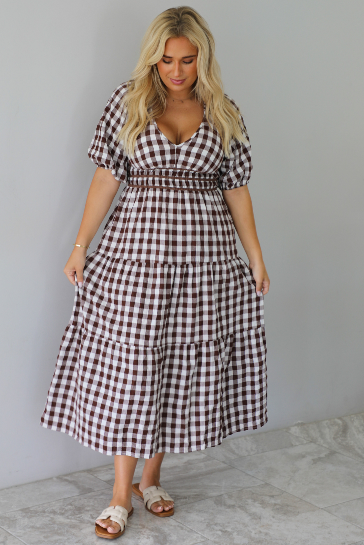 A woman modeling a brown and white gingham tiered midi dress with puff sleeves and a smocked waist, paired with beige slide sandals, standing indoors against a white wall with white marble flooring. 