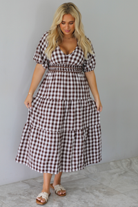 A woman modeling a brown and white gingham tiered midi dress with puff sleeves and a smocked waist, paired with beige slide sandals, standing indoors against a white wall with white marble flooring. 