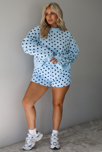 A woman wearing a blue two piece set with a navy polka dot pattern throughout, featuring a sweatshirt and high waisted sweat-shorts, paired with chunky sneakers and high ankle socks, shown indoors against a white wall with white marble flooring. 