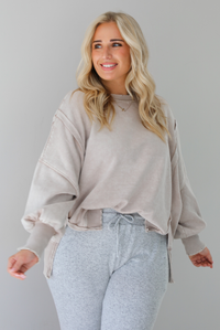 Woman wearing sand colored oversized sweatshirt with exposed seam details and a high-low design with heather grey casual pants. The photo is taken indoors against a white wall.