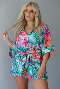 Woman wearing a multi-colored tropical romper with a collared neckline, flowy short sleeves, a cinched with a tie waistline, and flowy shorts indoors against a white wall.
