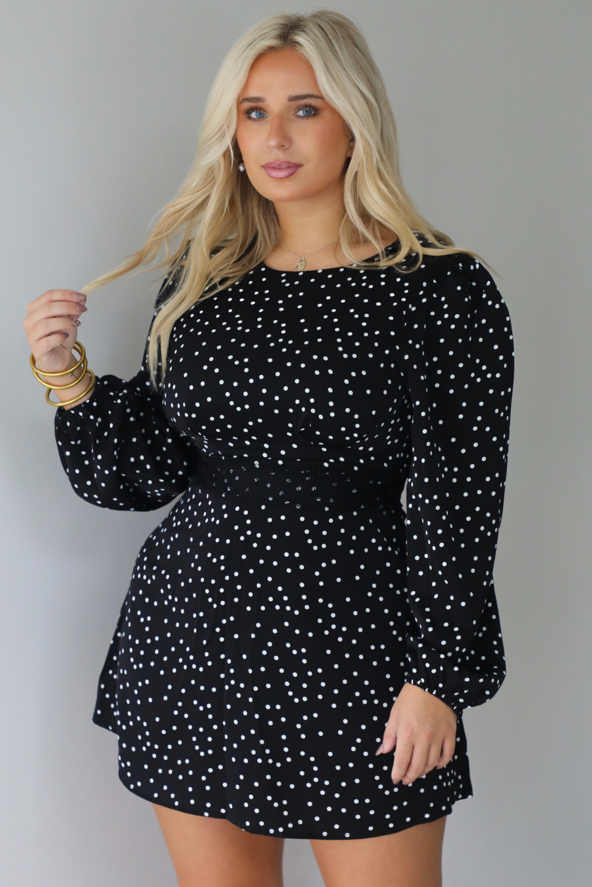 A woman wearing a black mini dress with a white polka dot print pattern throughout that has long sleeves, a high neckline, and a flowy skirt, standing indoors against a white wall.