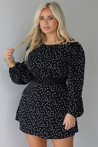 A woman wearing a black mini dress with a white polka dot print pattern throughout that has long sleeves, a high neckline, and a flowy skirt, standing indoors against a white wall.