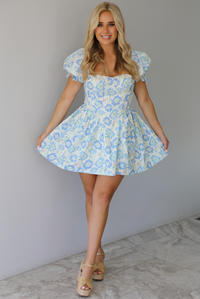 Woman wearing a white, blue, and green floral print mini dress that has short, puffed sleeves, a fitted corset bodice, and a flowy skirt indoors against a white wall with white marble flooring. 