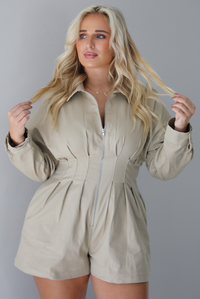 A woman wearing a sand colored long sleeved romper with a collared neckline, functional zipper closure in the front, pleated details throughout, functional pockets on either side, standing indoors against a white wall.
