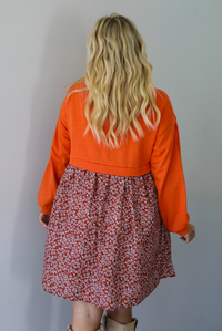 A woman shown from behind wearing a block style oversized, flowy dress featuring a solid orange bodice and sleeves with a multi-colored floral skirt, standing indoors against a white wall.