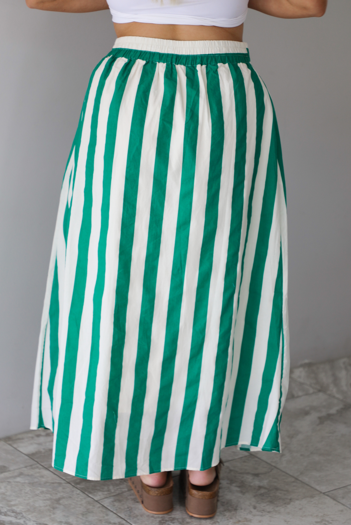 Closeup of the back of a woman wearing a white tank top and a maxi length cream and green striped skirt, focused in on the skirt, which has a high waistline, functional pockets on either side, functional button closures in the front, and has a flowy fit, standing indoors against a white wall with white marble flooring. 
