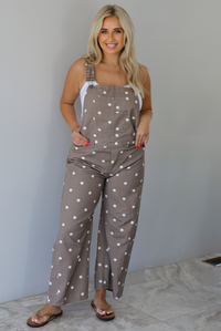 Woman wearing a brown with white polka dots overalls jumpsuit indoors against a white wall with white marble flooring. 