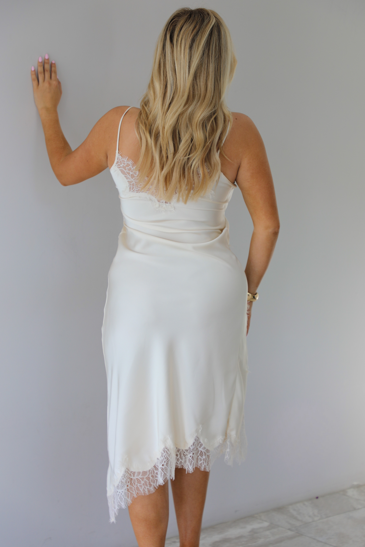 Woman wearing an off white satin midi length dress that has spaghetti straps, lace detailing at the v-neckline, and lace detailing at the hem and split hem detail on the right side. The photo is taken indoors against a white wall and shows the back of the dress.